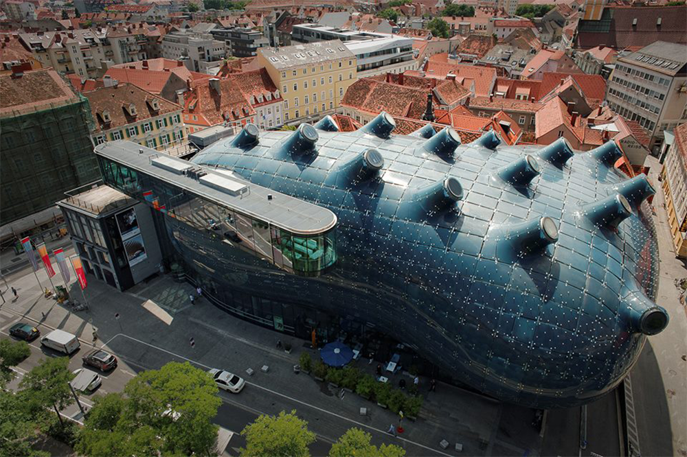 Aerial view of a large, dark blue, blob-shaped modern building with rounded protrusions, surrounded by traditional red-roofed city buildings and streets.
