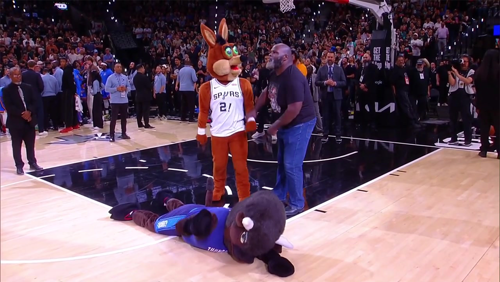 A man and a San Antonio Spurs mascot stand on a basketball court while an Oklahoma City Thunder mascot lies face down on the floor, surrounded by a crowd.