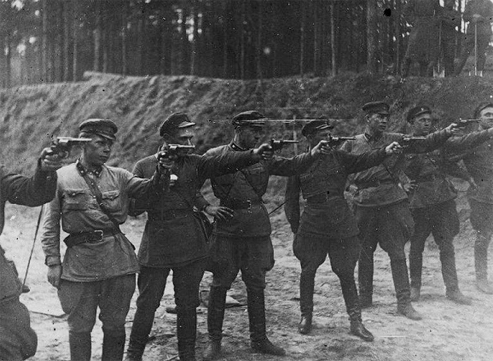 A group of uniformed soldiers stands outdoors, aiming pistols forward. The background features a dirt embankment and trees, suggesting a forested area. The atmosphere appears tense and somber.