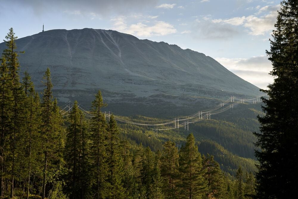 Power lines run through a forested valley with a large, rocky mountain in the background under a partly cloudy sky.