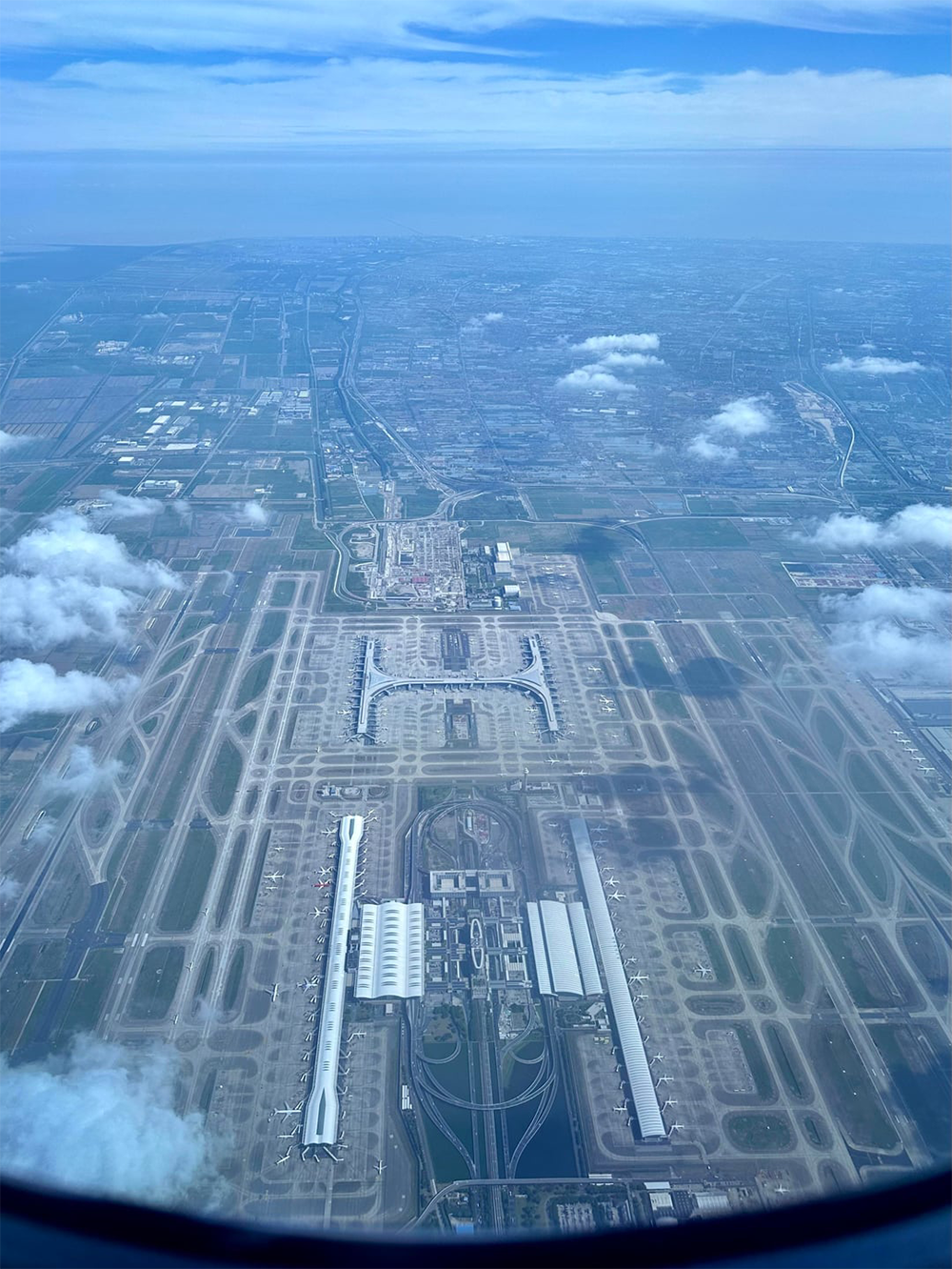 Aerial view of a large airport with multiple runways, taxiways, and terminals, seen through an airplane window with clouds and cityscape in the background.