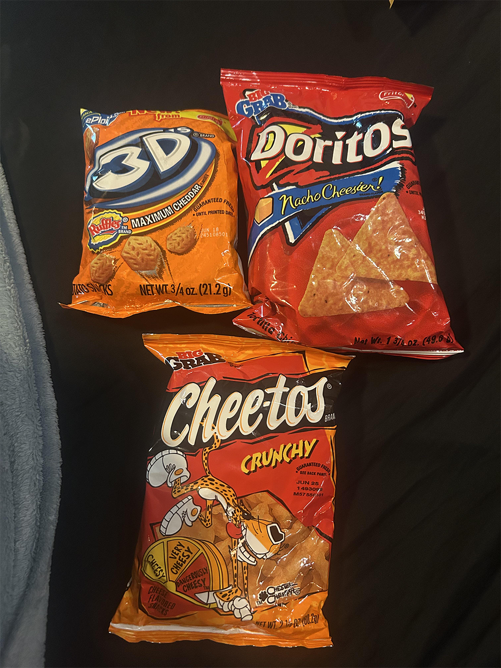 Three snack bags on a black surface: Doritos 3D, Doritos Nacho Cheese, and Crunchy Cheetos. Each bag is unopened and brightly colored with images of the snacks on the packaging.