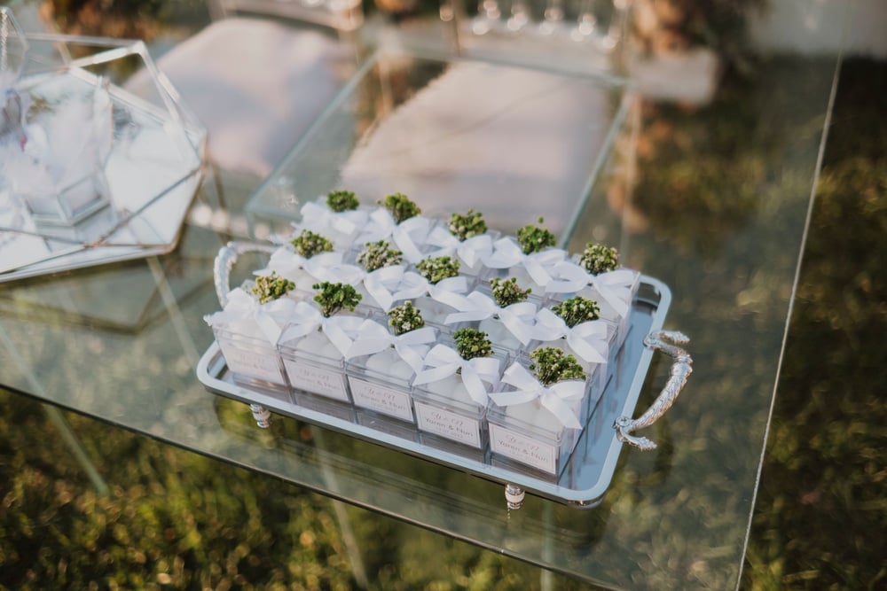 A glass table holds a tray of small white gift boxes, each topped with a sprig of greenery and a white ribbon, arranged neatly for an event.