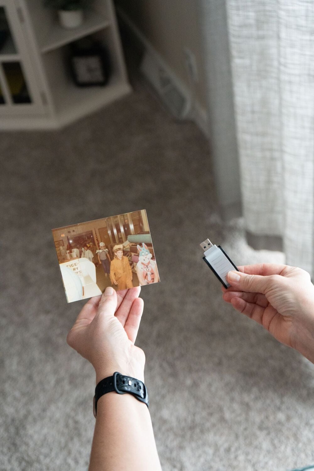 A person holds an old photograph in one hand and a USB flash drive in the other, standing over a carpeted floor near a window and a white cabinet.