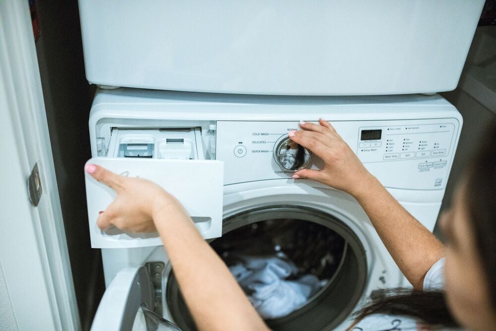 A person loads detergent into a front-loading washing machine, with one hand opening the detergent drawer and the other adjusting the control knob.