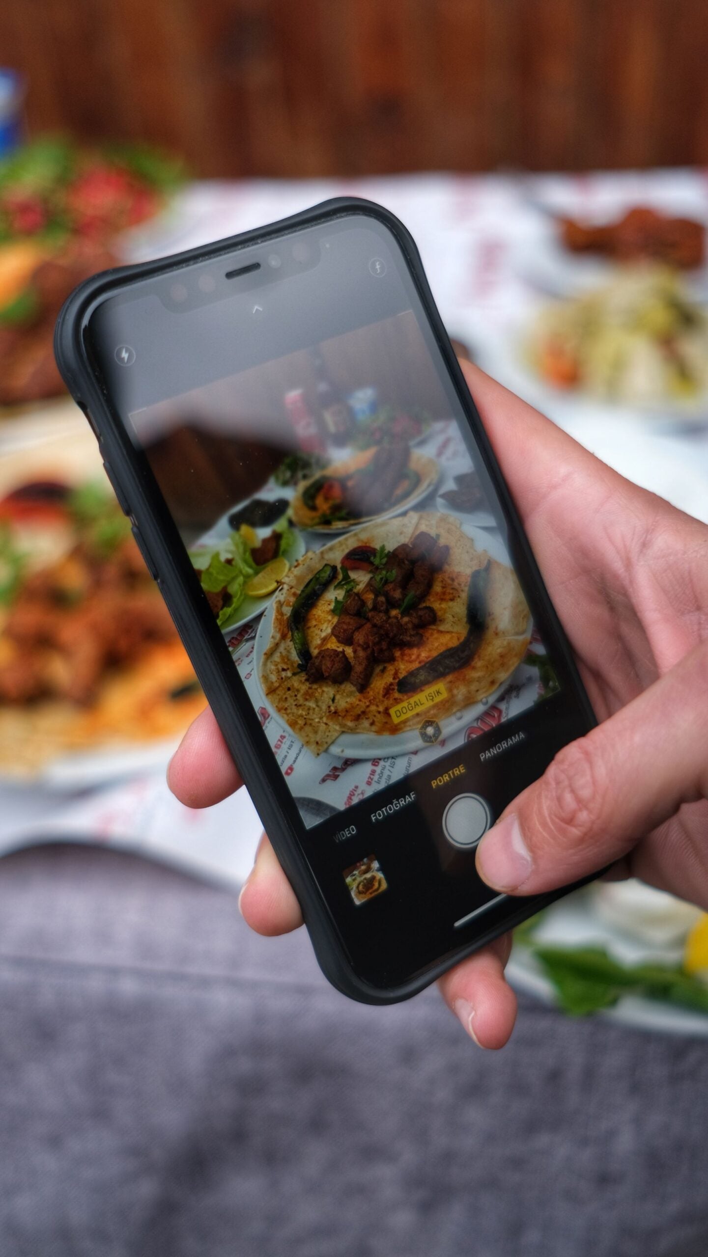 A hand holds a smartphone, taking a photo of a plate of food with meat, vegetables, and flatbread. The smartphone screen shows the focused dish, while the real food is slightly blurred in the background.