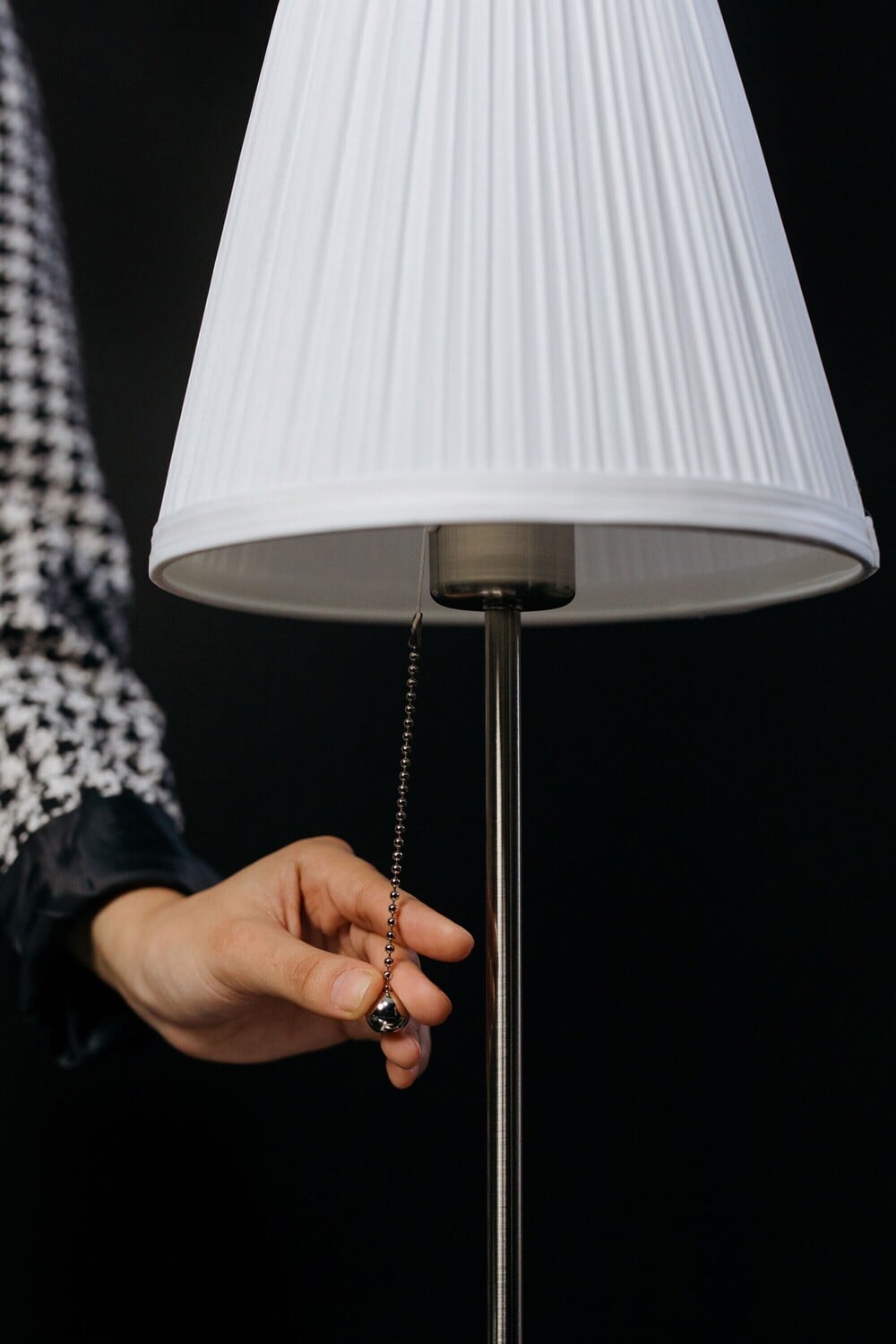 A person’s hand is pulling the chain of a standing lamp with a white pleated lampshade against a dark background. Only part of the person’s arm, dressed in a houndstooth sleeve, is visible.