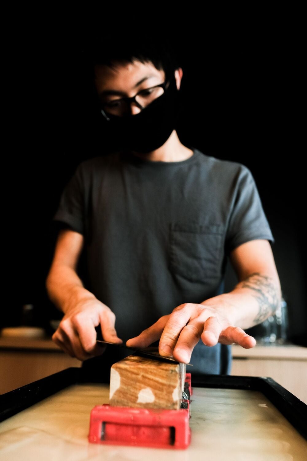 A person wearing glasses and a mask uses both hands to sharpen a knife on a whetstone, with a dark background and focus on their hands.