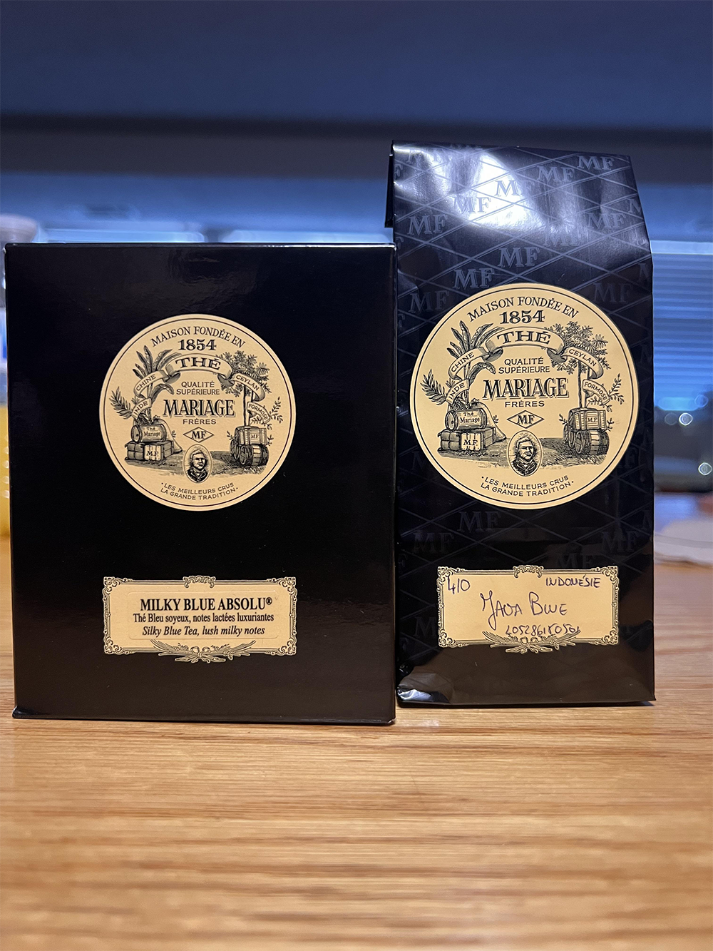 Two black Mariage Frères tea packages labeled “Milky Blue Absolu” are on a wooden surface. One is a box, the other a bag with handwritten details. Both feature a gold, vintage-style logo.