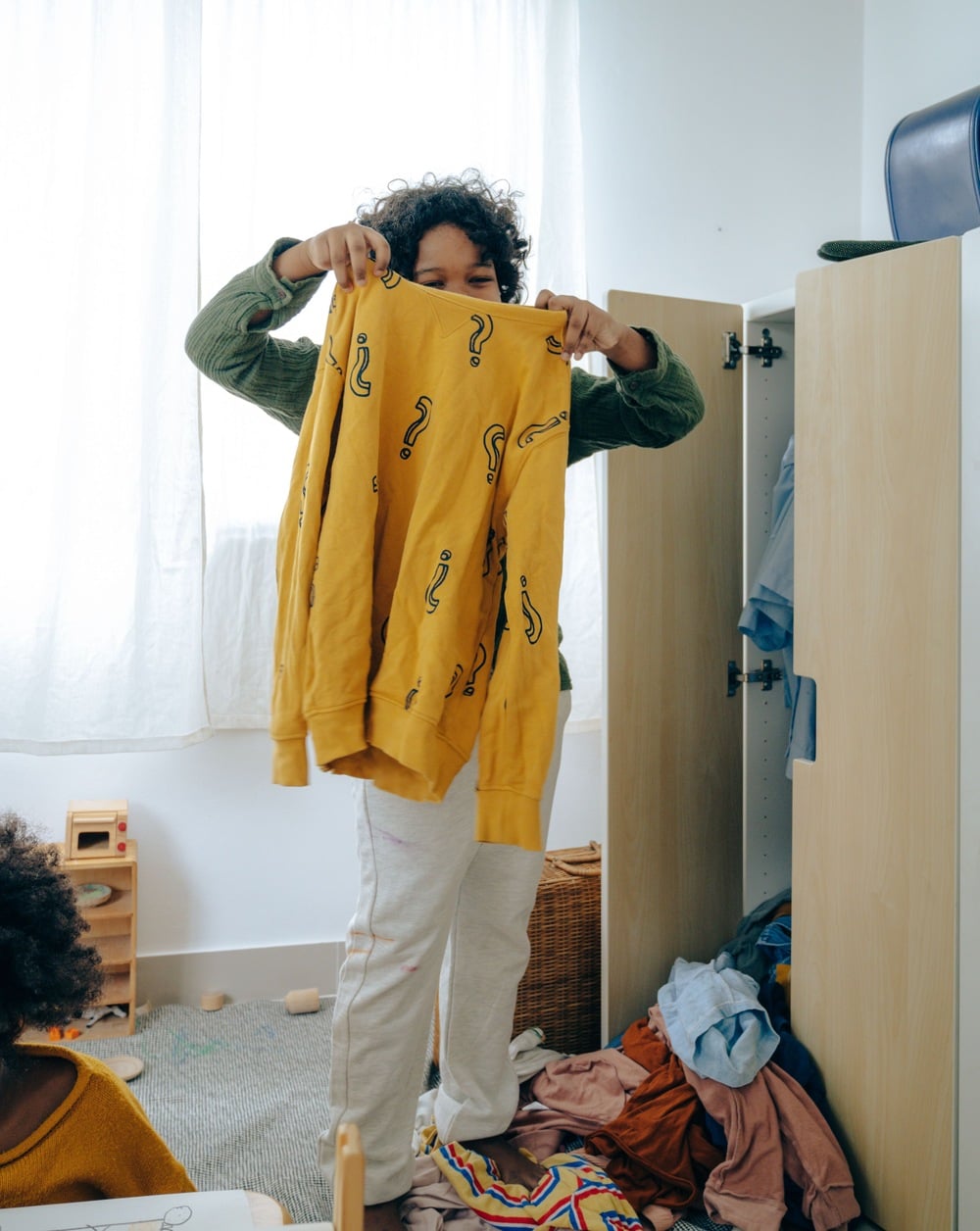 A child holding up a yellow sweatshirt with question marks, standing by an open wardrobe with clothes inside and more clothes scattered on the floor.