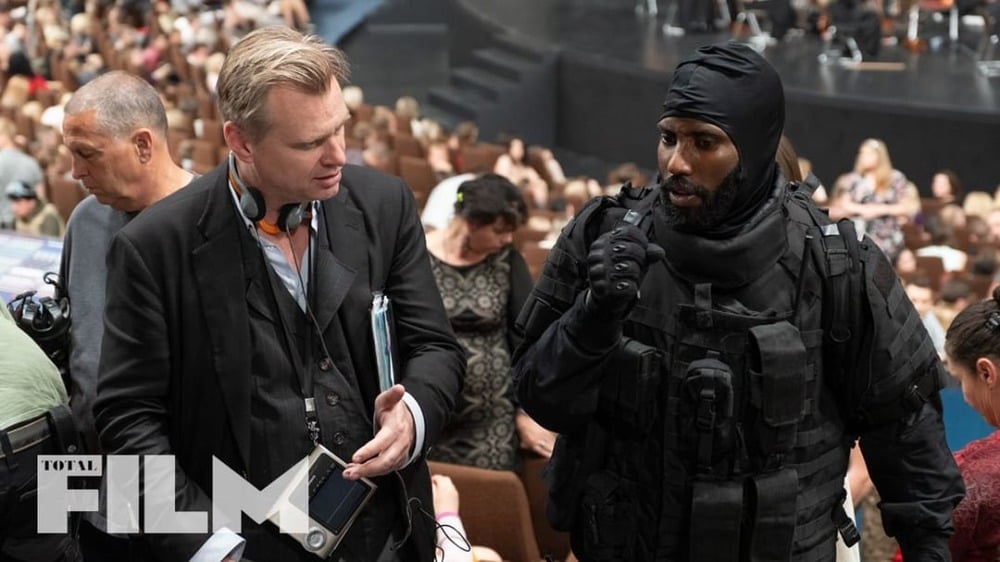 A man in a suit talks to a person in black tactical gear at an event with many seated people. The Total Film logo is visible in the corner.