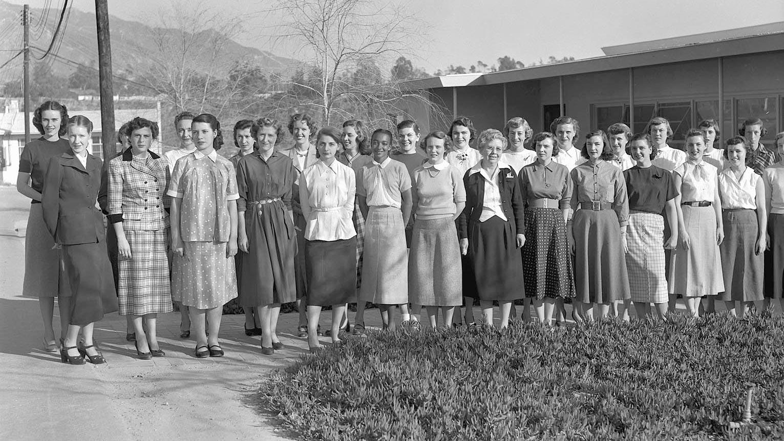 A group of women in mid-century clothing stand in a row outdoors, posing for a black-and-white photo in front of a building and some trees.