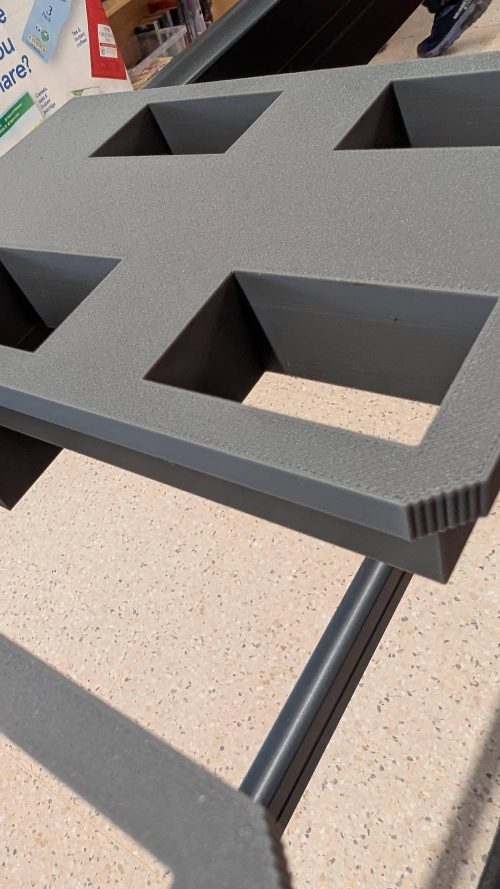 A close-up of a gray, textured, rectangular object with four square cutouts, resting on a metal frame. The background shows a speckled tile floor and some partially visible product packaging.