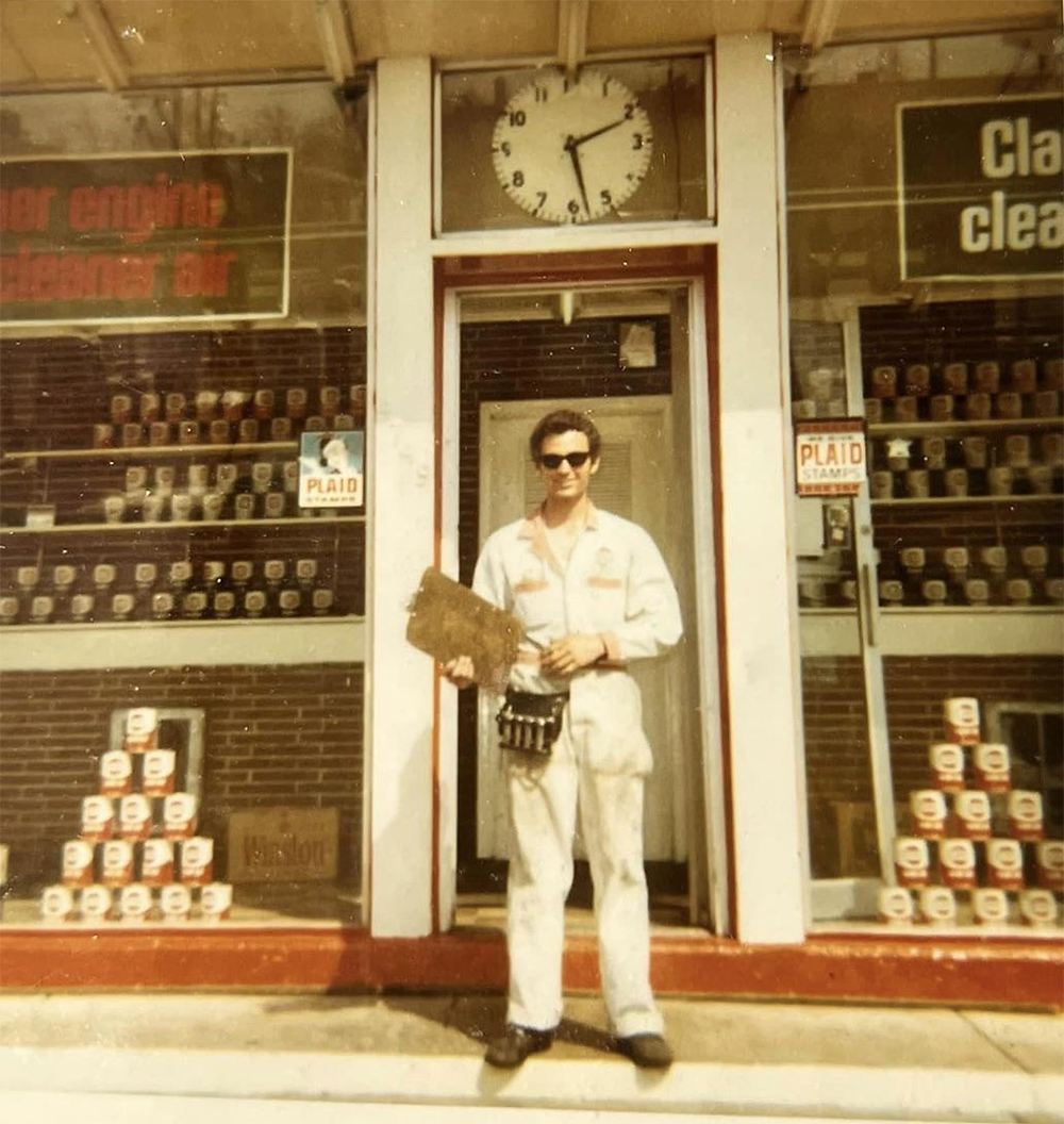 A person in a white uniform stands in front of a store with jars displayed in the windows. A clock above the door shows 12:15. The person holds a clipboard and wears sunglasses.