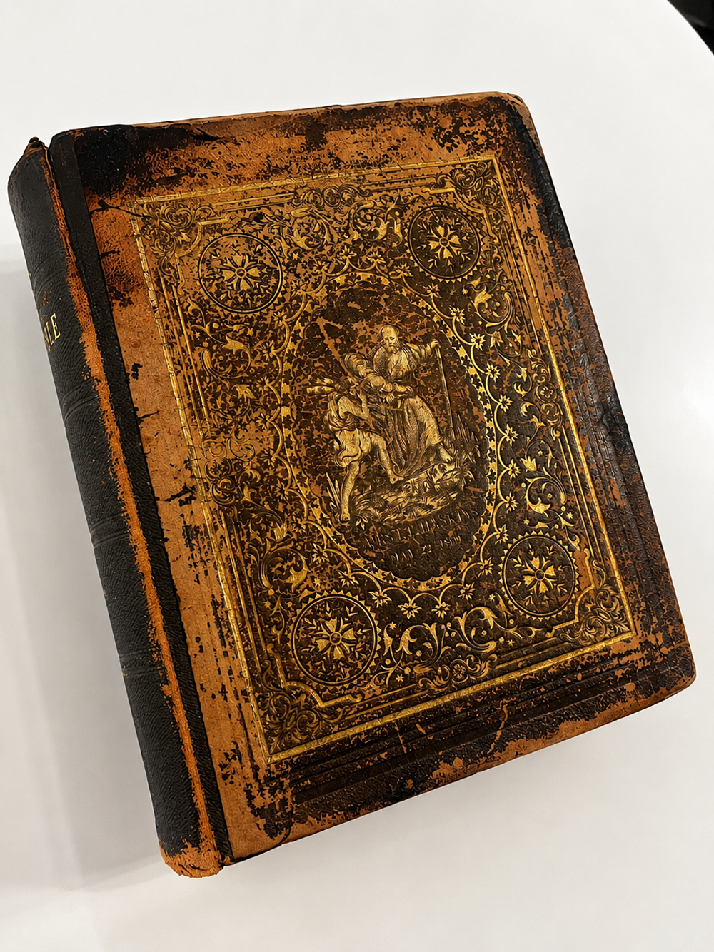 An old, ornate book with a worn brown leather cover featuring intricate gold detailing and an embossed illustration of two figures in the center.