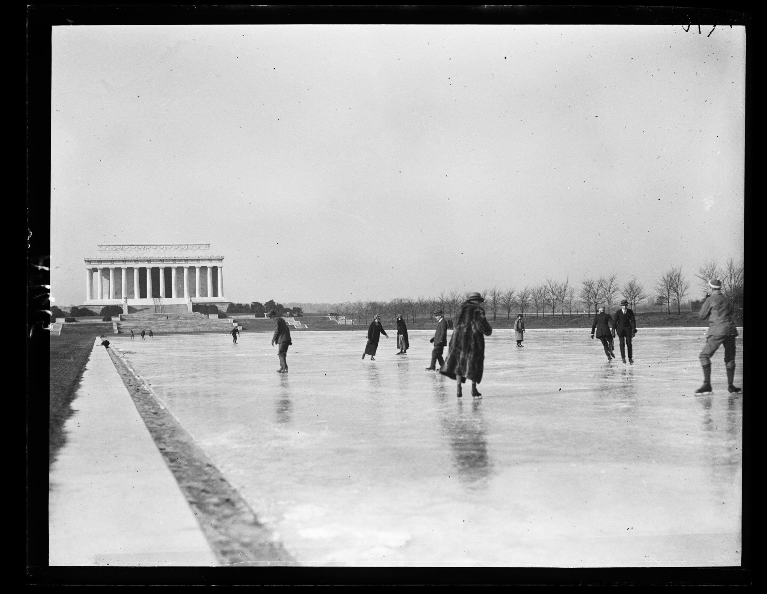 People ice skating on a frozen reflecting pool near the Lincoln Memorial in Washington, D.C., on a cloudy day. Leafless trees line the background.