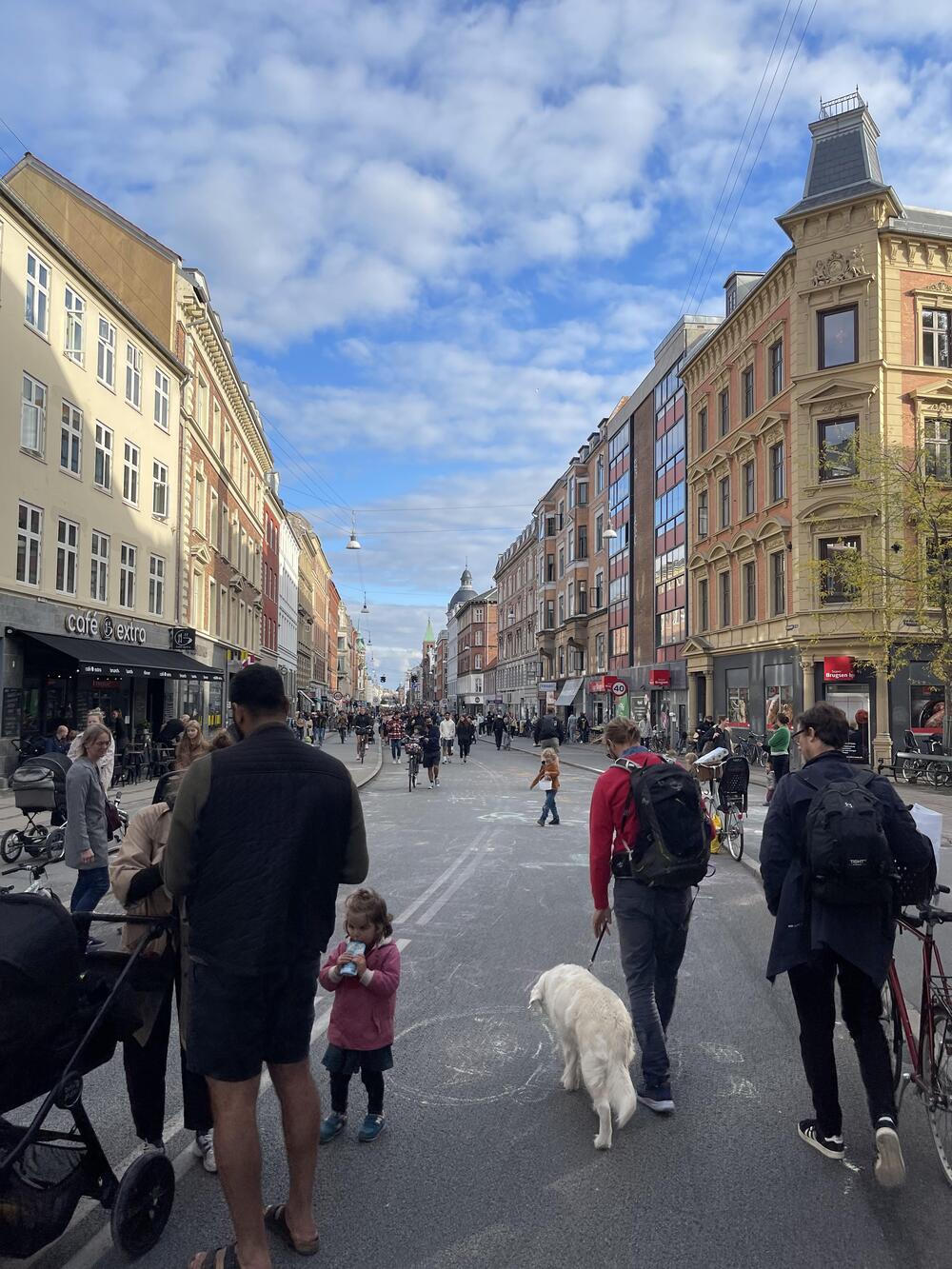A busy city street with people walking, cycling, children playing, and a dog. Buildings line both sides under a partly cloudy blue sky. Outdoor cafes and shops are visible along the sidewalk.