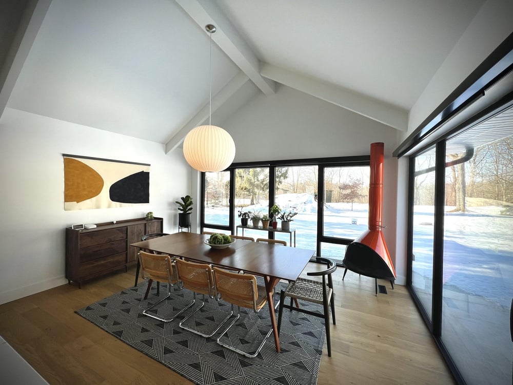 A modern dining room with wooden table, six chairs, geometric rug, pendant light, sideboard, wall art, plants, orange fireplace, and large windows showing a snowy yard.