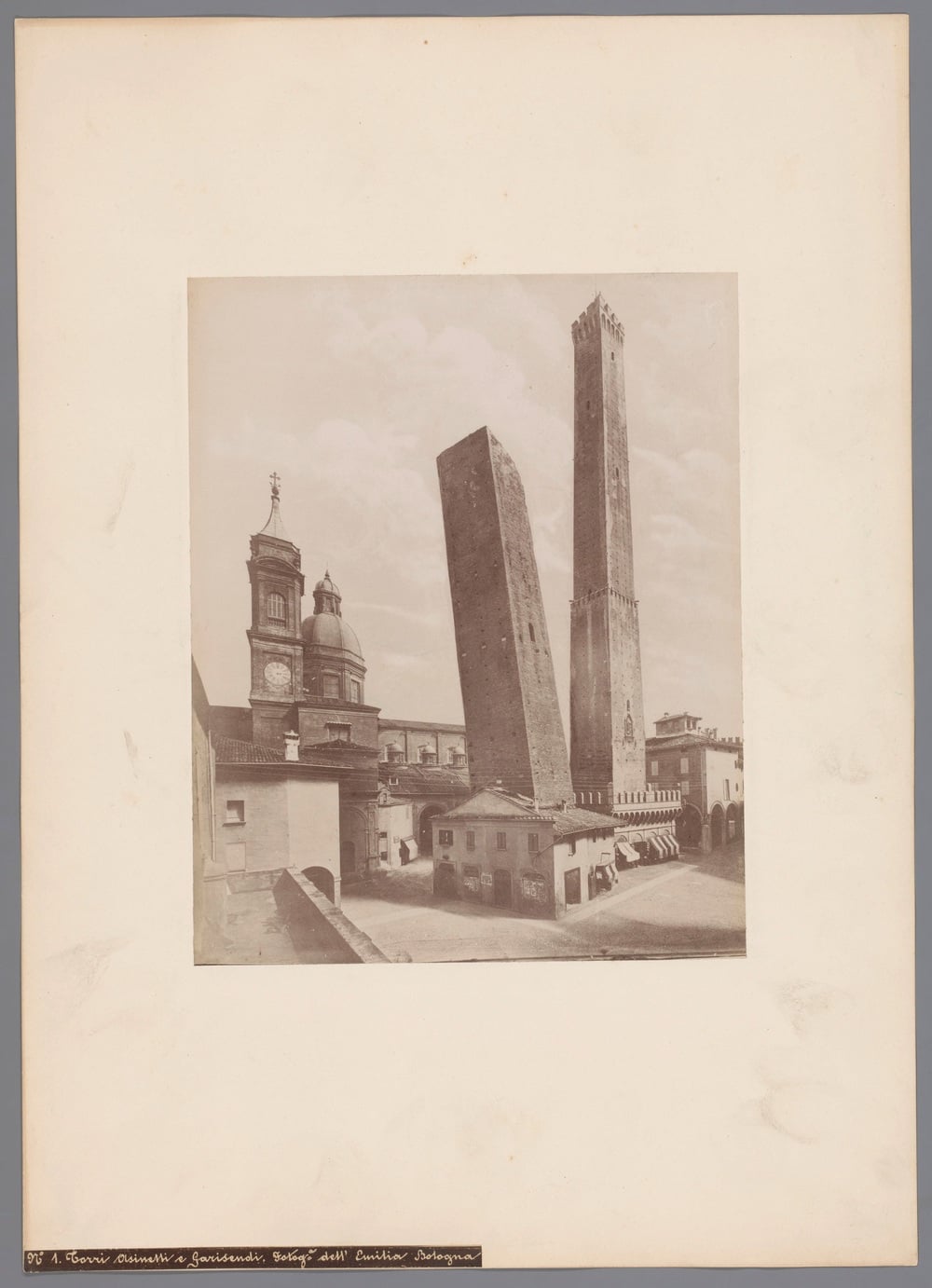 Historic black-and-white photo of Bologna’s iconic Two Towers—Asinelli and Garisenda—standing next to old buildings and a church with domes, under a clear sky.