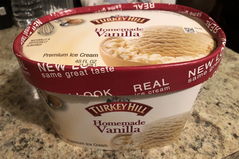 A 48 oz container of Turkey Hill Homemade Vanilla Premium Ice Cream sits on a granite countertop. The lid highlights “New Look” and “Real Ice Cream.” The label shows an image of vanilla ice cream.
