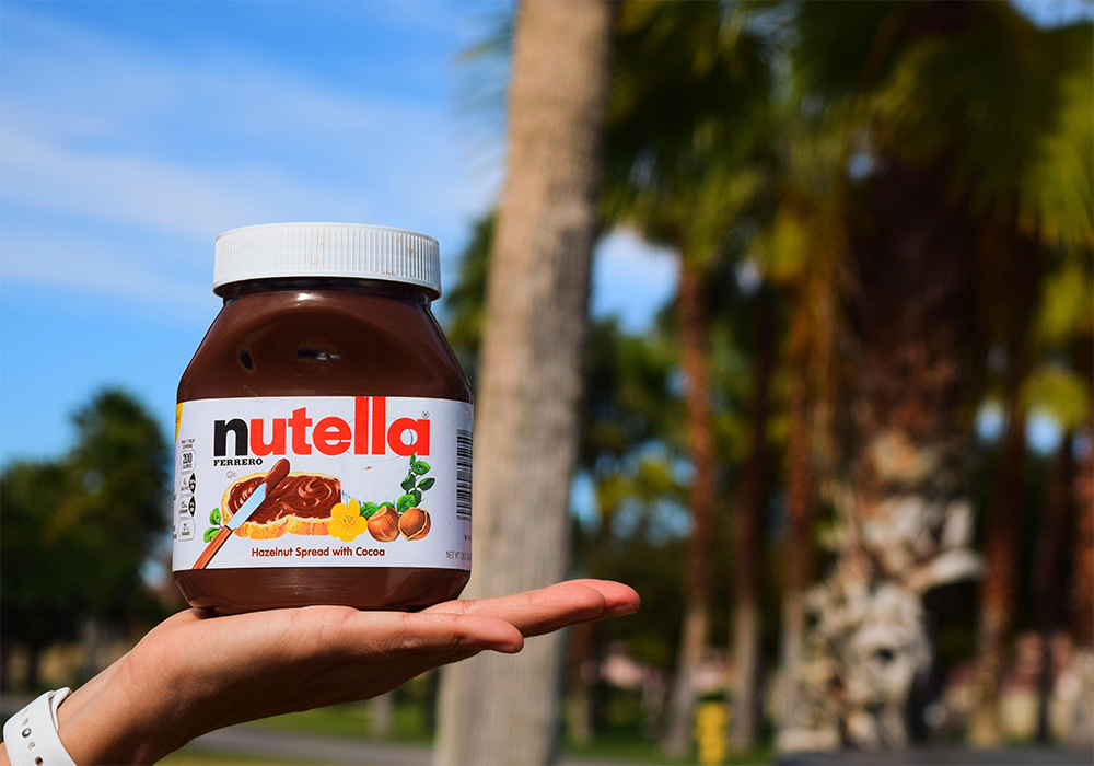 A hand holding a large jar of Nutella hazelnut spread outdoors, with blurred palm trees and greenery in the background on a sunny day.