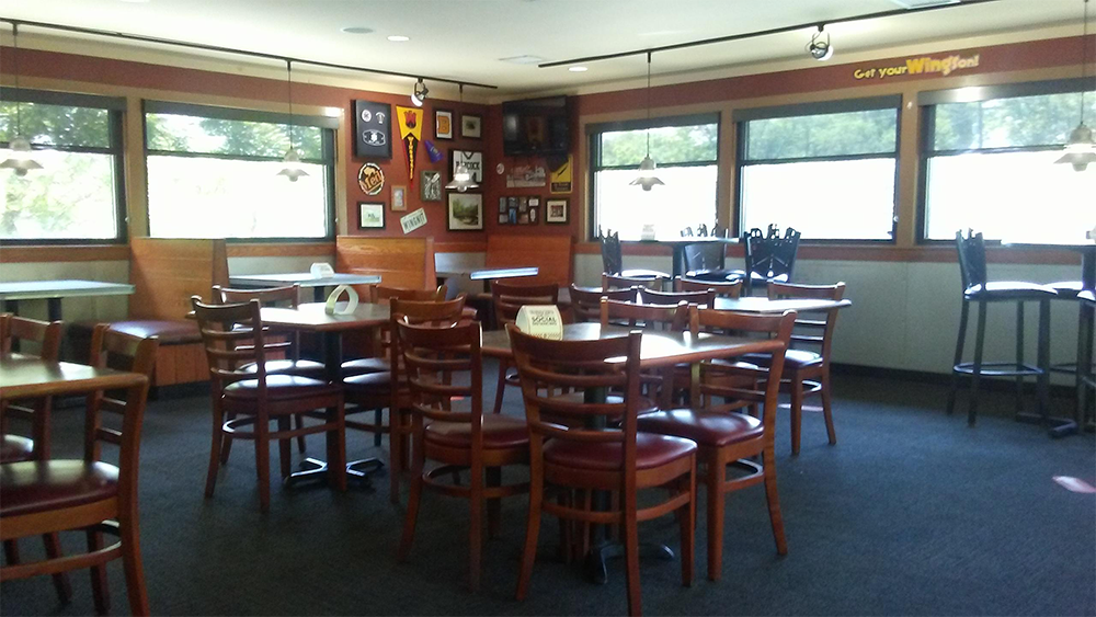 A casual restaurant dining area with wooden tables and chairs, large windows letting in sunlight, and sports decor on the walls. The space is empty and well-lit, with a mix of booth and table seating.