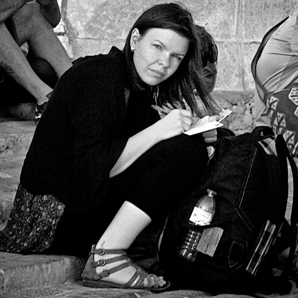 A woman sits on stone steps, writing in a notebook. She wears sandals and a dark outfit, with a backpack and water bottle beside her. The image is black and white.