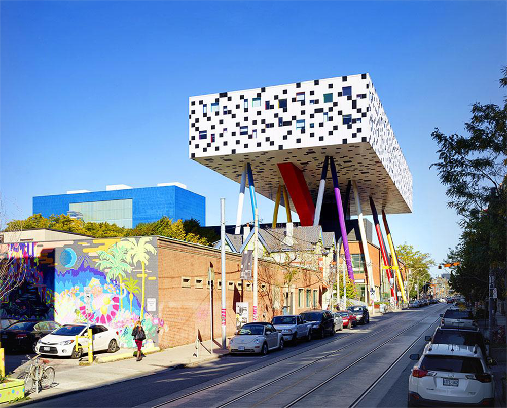 A modern building with a white, pixel-patterned upper section on colorful stilts towers over a street lined with cars, murals, and pedestrians on a sunny day.