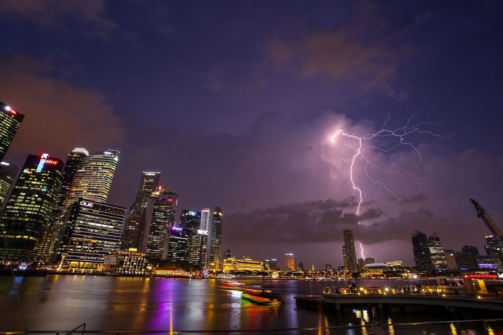 A city skyline at night with tall buildings lit up, a river reflecting colorful lights, and a bright lightning bolt illuminating the cloudy sky.