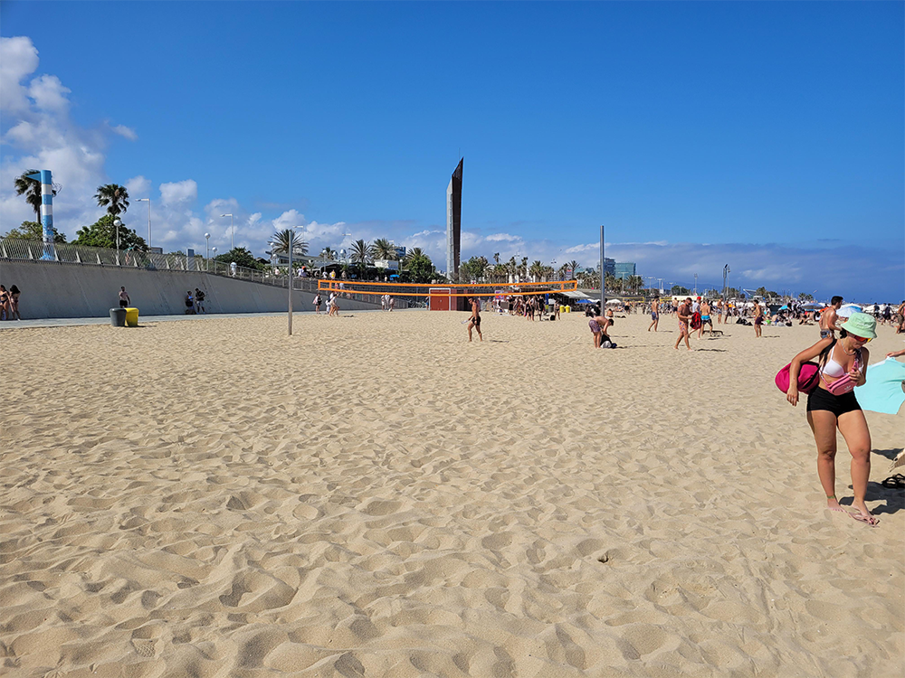 People enjoy a sunny day at a sandy beach with a blue sky, some clouds, and palm trees. A few structures and a tall sculpture are visible in the background.