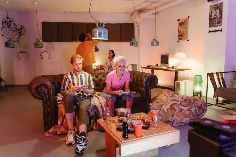 Two people sit on a brown couch playing video games, snacks and drinks on a table in front of them. Two others stand in the background, talking. The room is decorated with quirky art and vintage items.