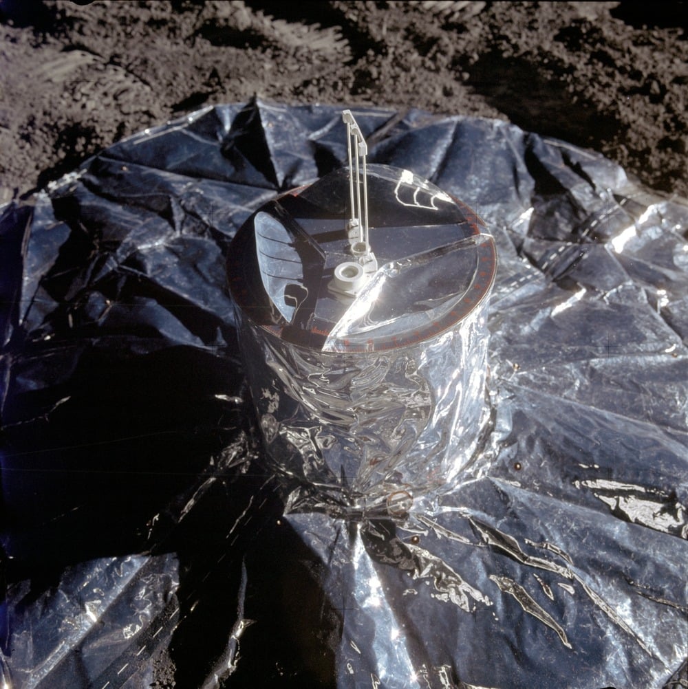 A shiny scientific instrument, possibly a seismometer, sits on a wrinkled metallic sheet on the moon’s surface, with lunar soil visible in the background.