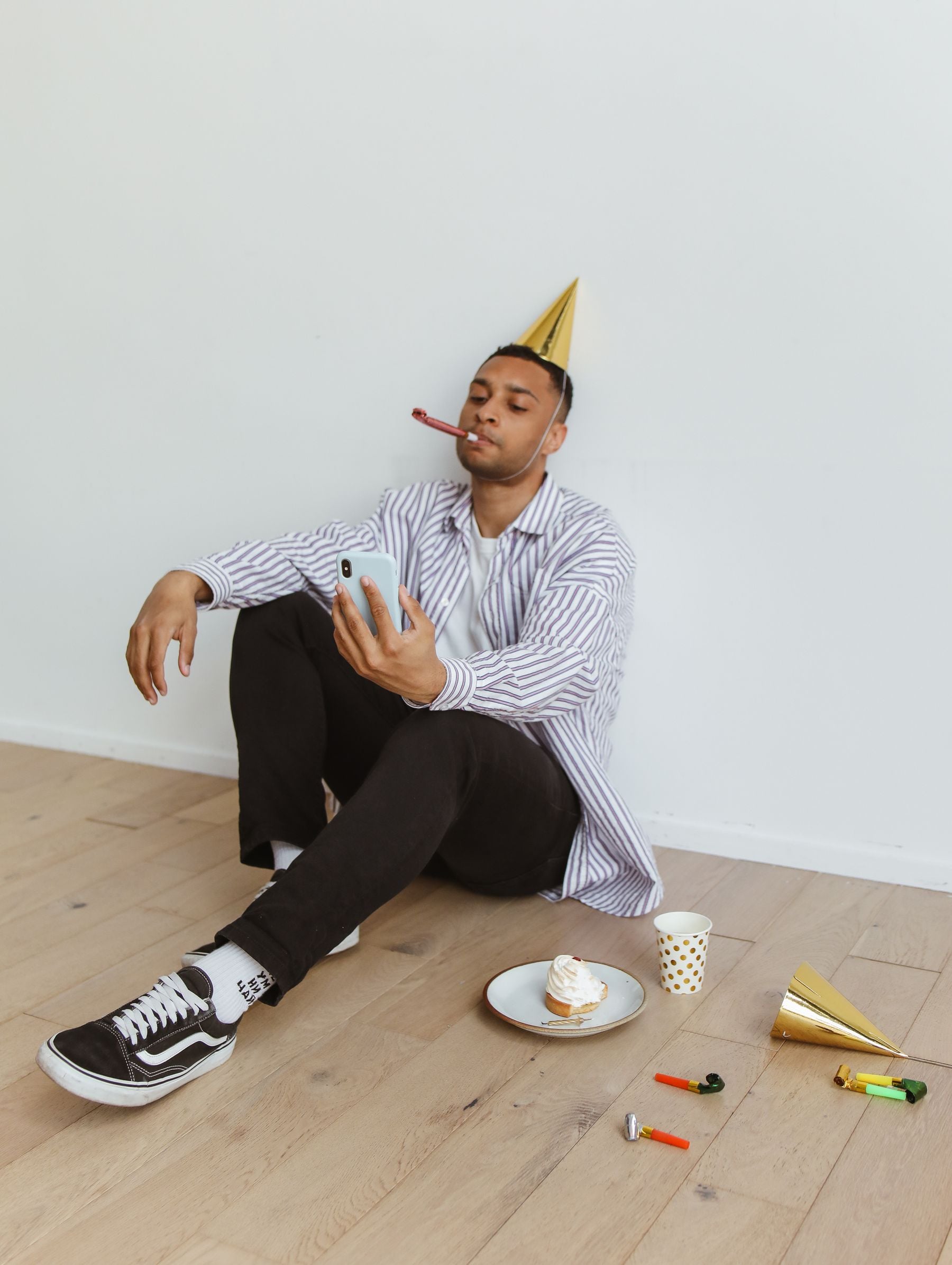 A person in a party hat sits on the floor, taking a selfie with their phone. There are party items, a cupcake, and an empty plate beside them. They appear relaxed and are blowing a party horn.