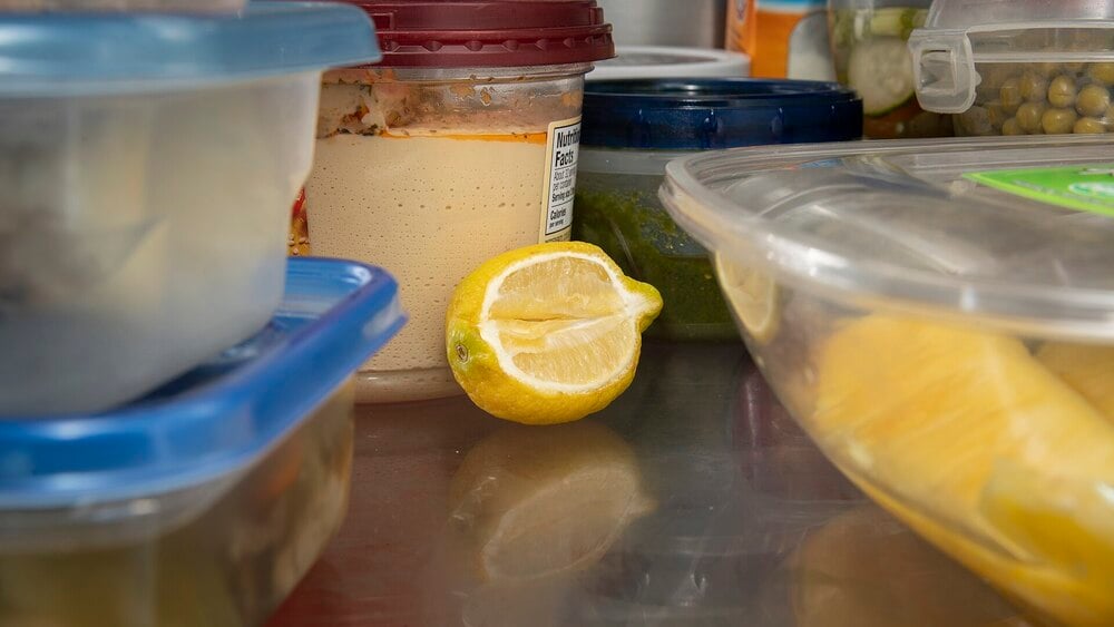 A half lemon sits on a refrigerator shelf surrounded by various plastic containers of food, some with clear and some with colored lids.