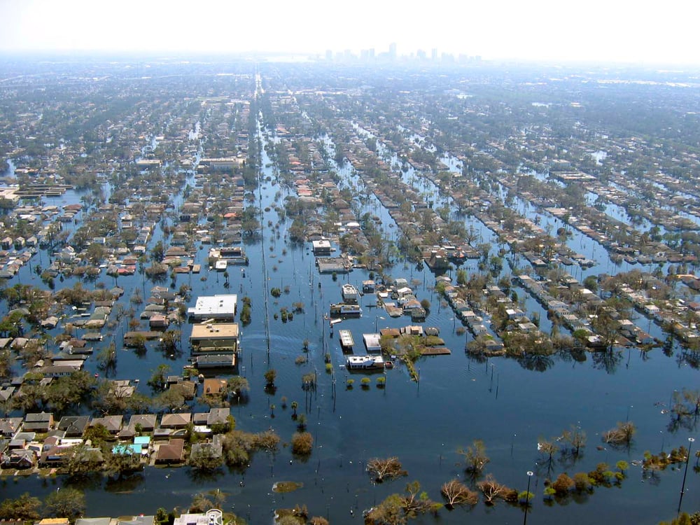 Aerial view of a flooded city neighborhood, with water covering streets and surrounding homes and buildings; distant city skyline visible in the background.