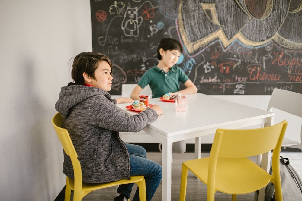 Two boys sit at a white table with yellow chairs, eating snacks. Behind them is a blackboard covered in colorful chalk drawings and writing.