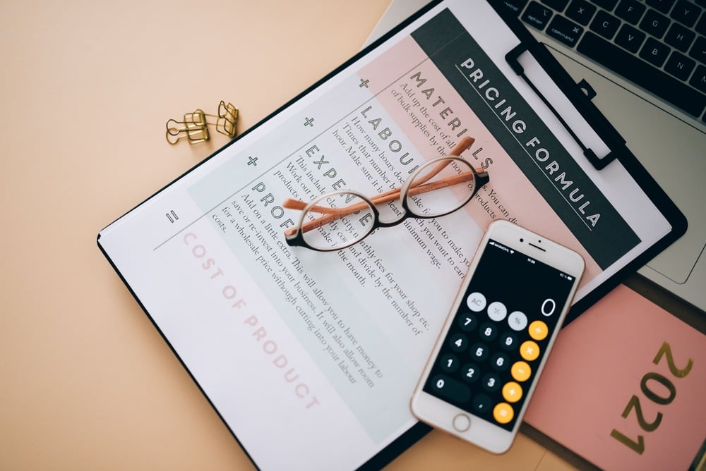 A clipboard with a pricing formula sheet, a pair of glasses, a smartphone with a calculator app open, and part of a laptop on a desk. A pink “2021” notepad and gold paper clips are also visible.