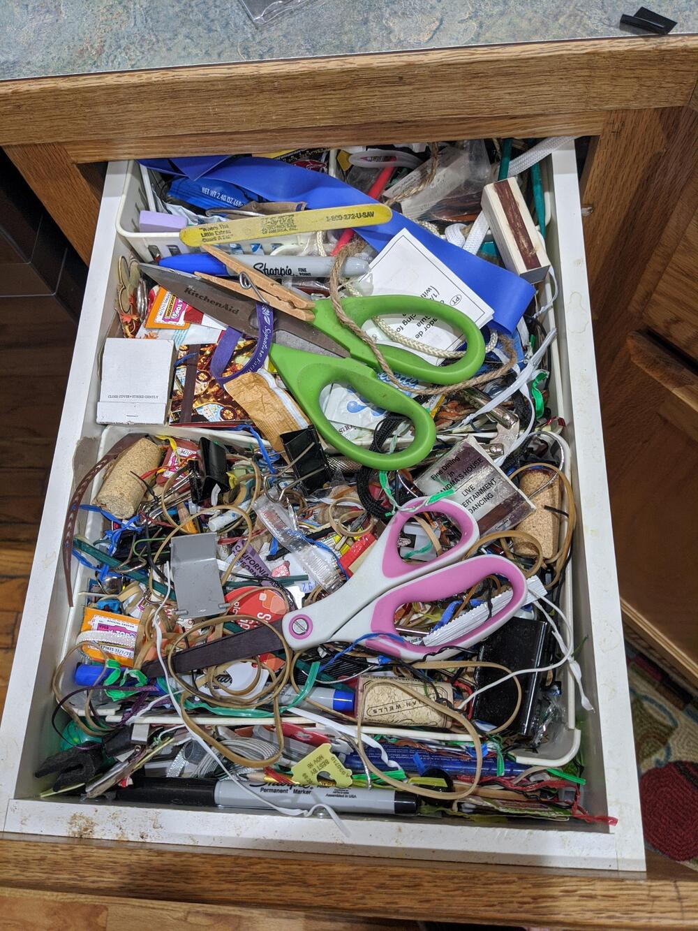 A messy drawer filled with various items, including two pairs of scissors, pens, cables, batteries, rubber bands, and miscellaneous household objects.