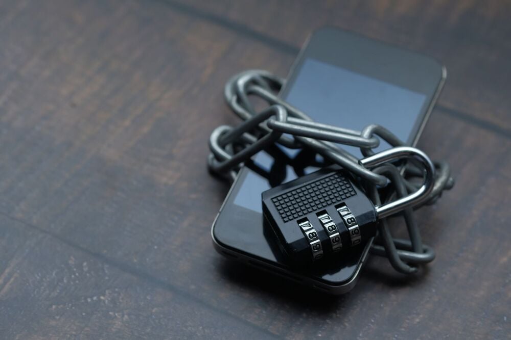 A smartphone wrapped in a metal chain and secured with a combination padlock, symbolizing data protection or restricted access, placed on a dark wooden surface.