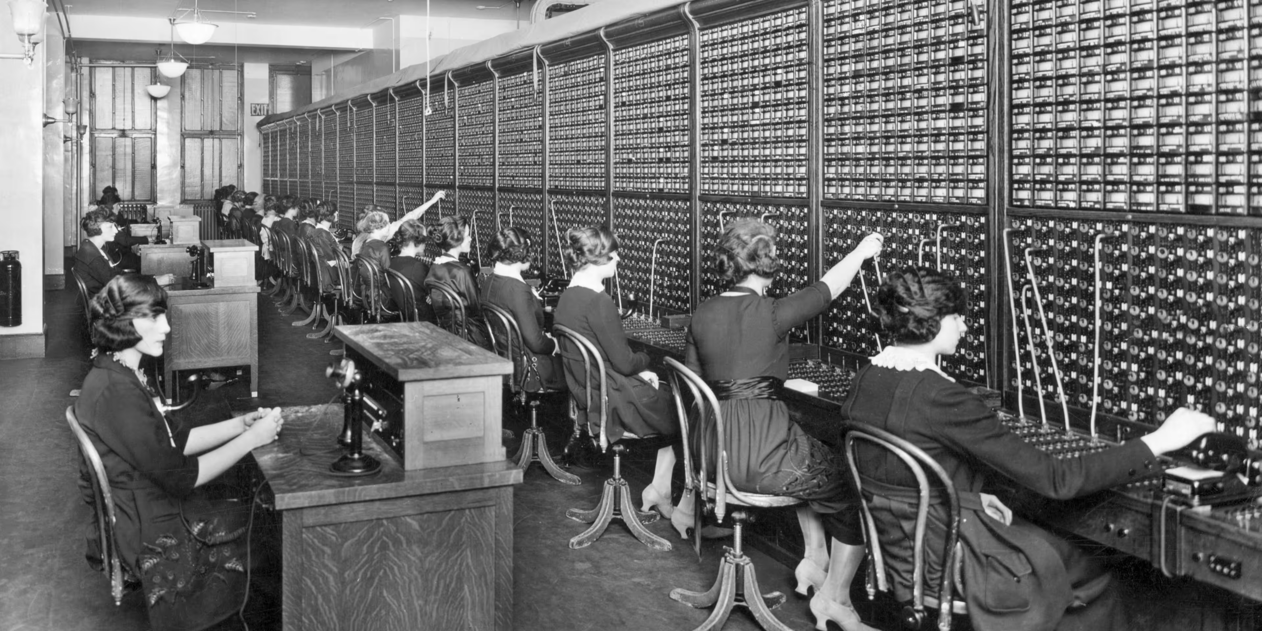 Rows of women operate a large, old-fashioned telephone switchboard, connecting calls by plugging in cables. They wear dark dresses and sit on chairs facing the switchboard in a busy, vintage office setting.