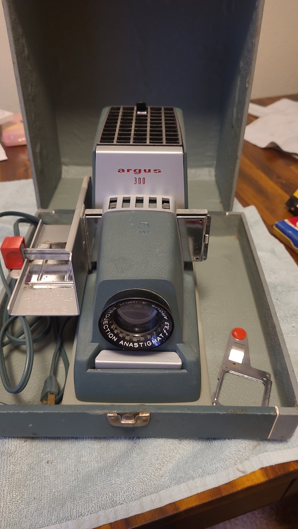 An Argus 300 slide projector sits in an open carrying case with its lens facing forward, power cord, and slide holder visible. The case rests on a light blue towel on a wooden table.