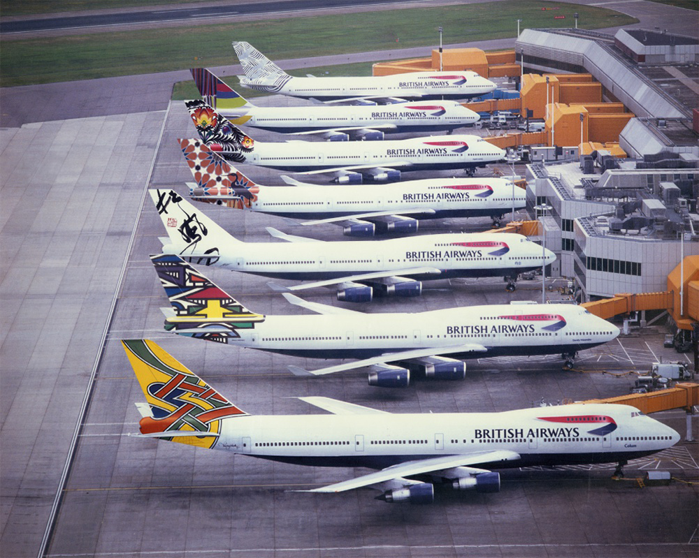Several British Airways planes with colorful tail designs are parked in a row at gates at an airport terminal, viewed from above. The planes have white bodies with blue underbellies and red accents.