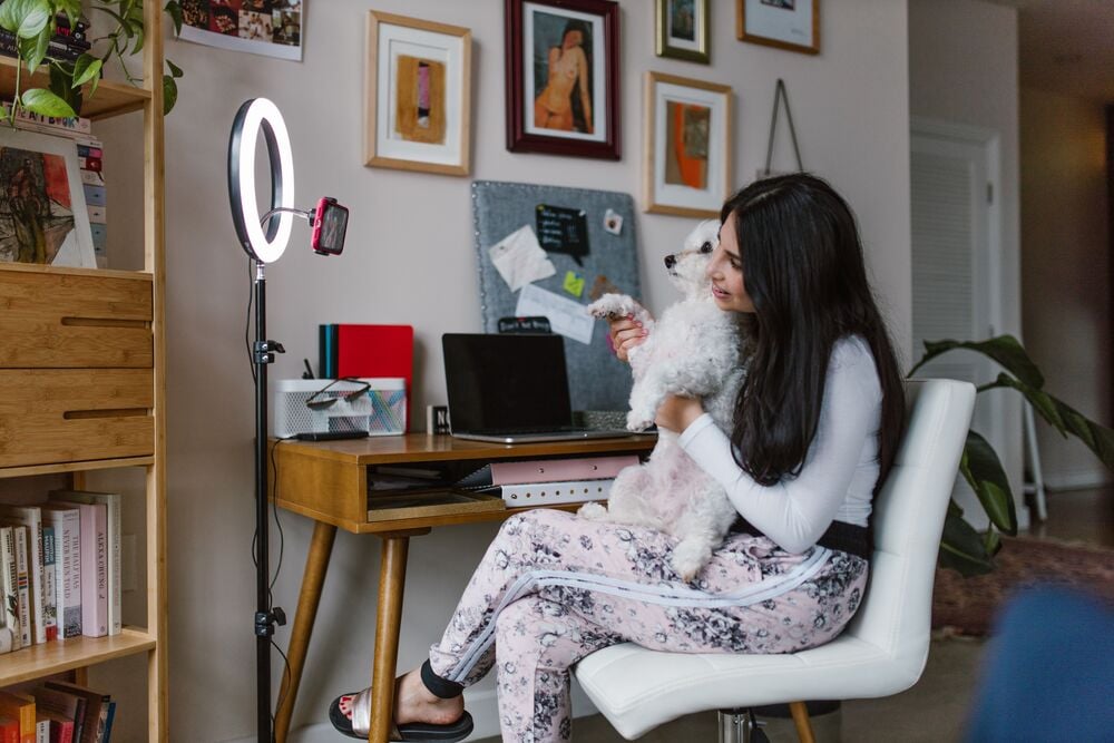 A woman sits at a desk in a home office, holding a small white dog and smiling. A ring light and phone are set up nearby, with shelves, books, and framed art on the wall behind her.