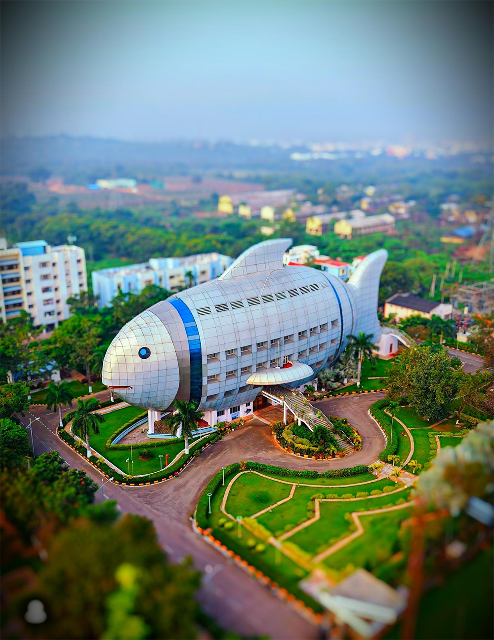 Aerial view of a large building shaped like a fish, surrounded by green lawns, palm trees, and nearby structures, set within a landscaped campus.