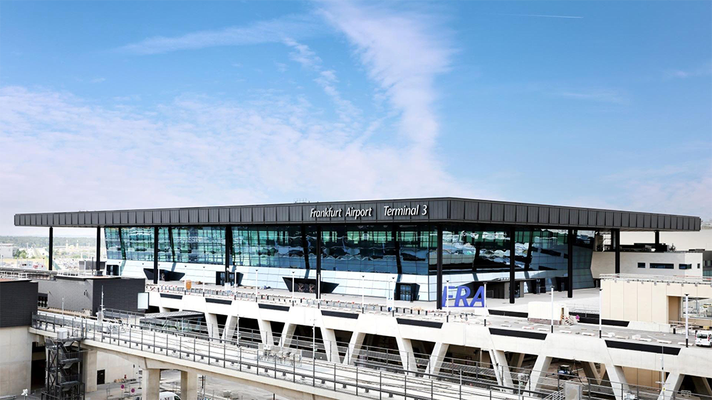 Frankfurt Airport Terminal 3 building with large glass windows, modern design, and visible tracks in the foreground under a clear blue sky.
