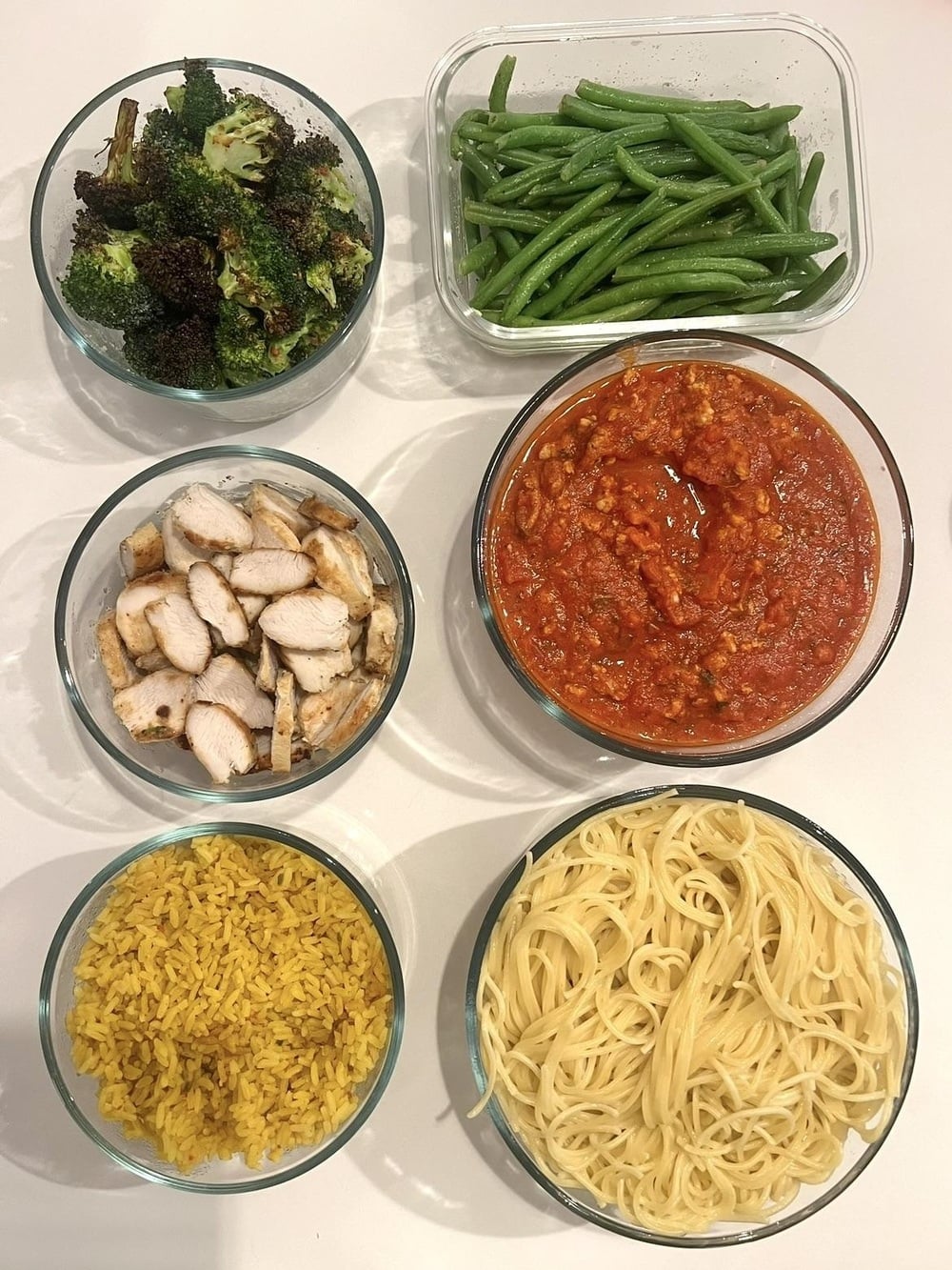 Six glass bowls with cooked foods: roasted broccoli, green beans, sliced grilled chicken, marinara sauce with ground meat, yellow rice, and cooked pasta, all arranged on a white countertop.
