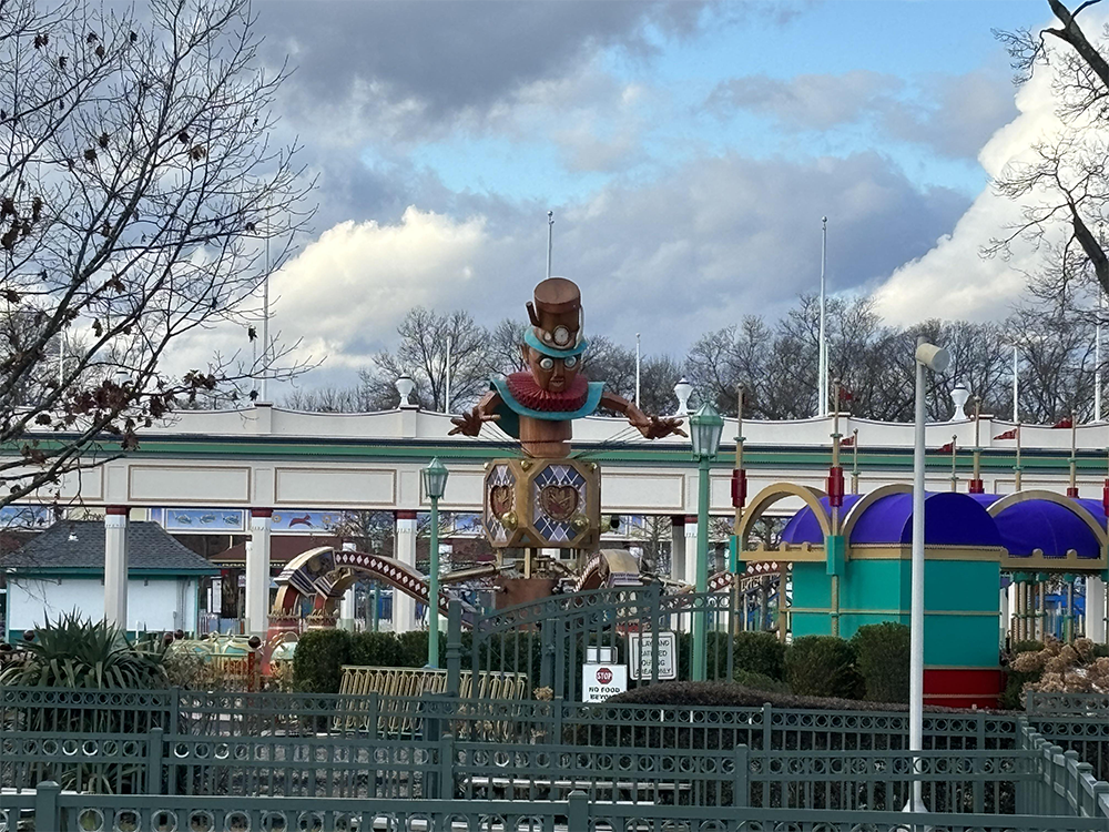 A large wooden statue of a smiling character in a hat and scarf stands in front of a colorful amusement park ride, with bare trees and cloudy skies in the background. A metal fence is visible in the foreground.