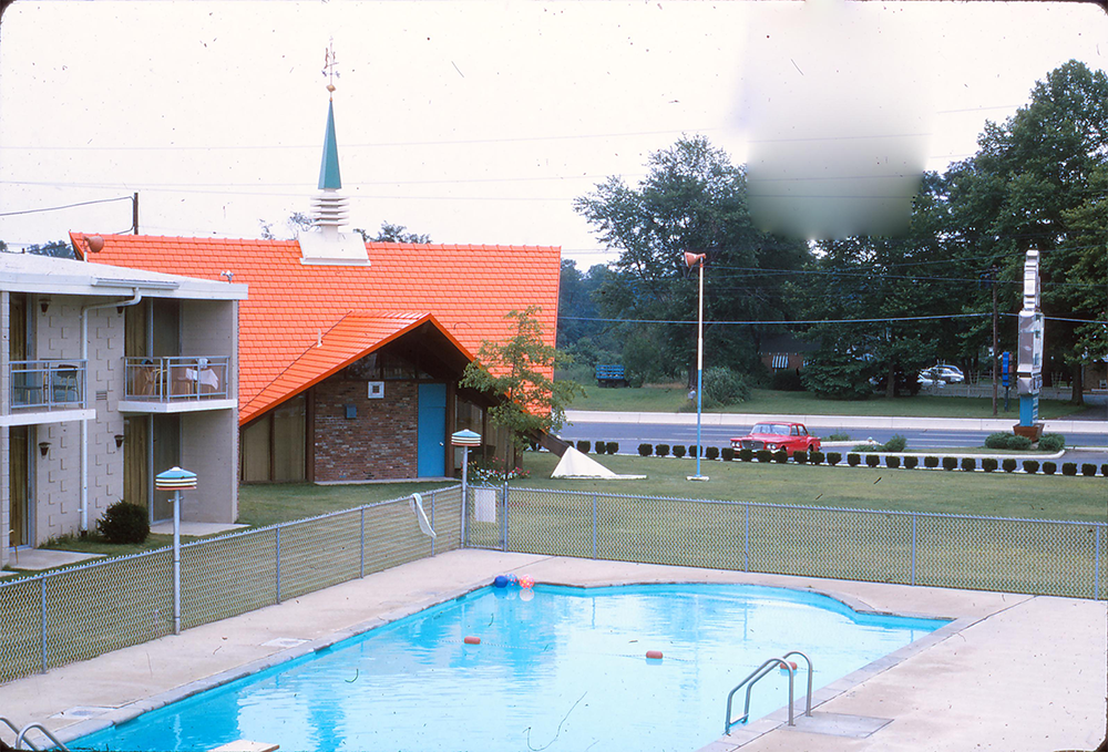 A swimming pool is in the foreground, fenced in beside a motel. Behind it is a small building with an orange roof and steeple, a grassy lawn, and a road with a red car and trees in the background.