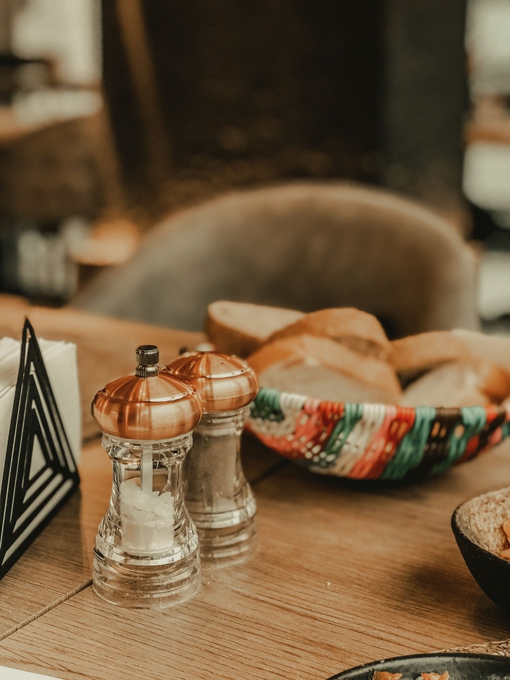 A close-up of salt and pepper shakers on a wooden table, with a colorful bowl filled with sliced bread in the background.