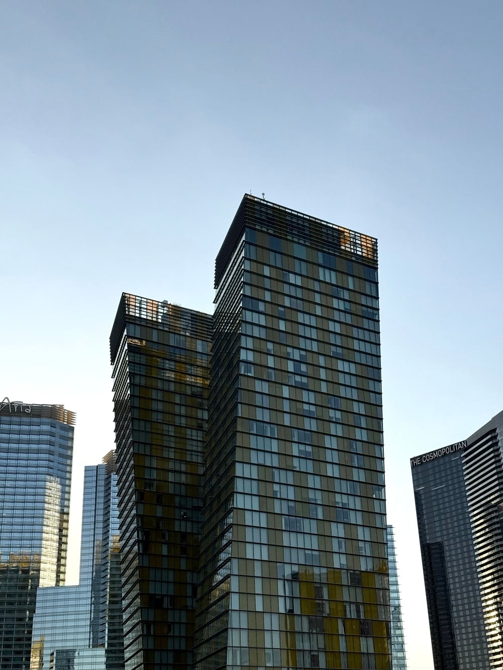Two modern high-rise buildings with reflective glass windows stand against a clear blue sky. Other tall buildings, including one labeled “The Cosmopolitan,” are visible in the background.